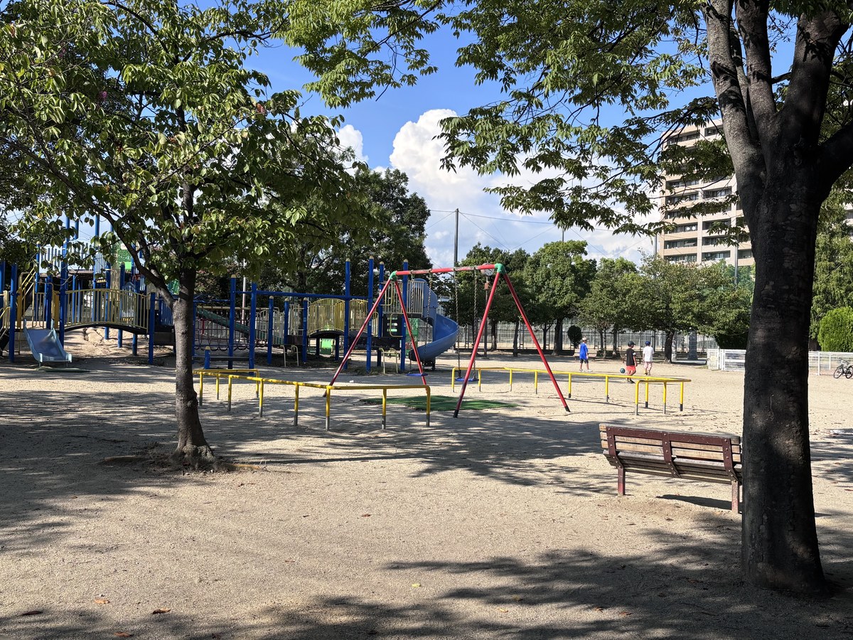 新高中央公園の風景。木陰のベンチの向こうに遊具とブランコが並ぶ